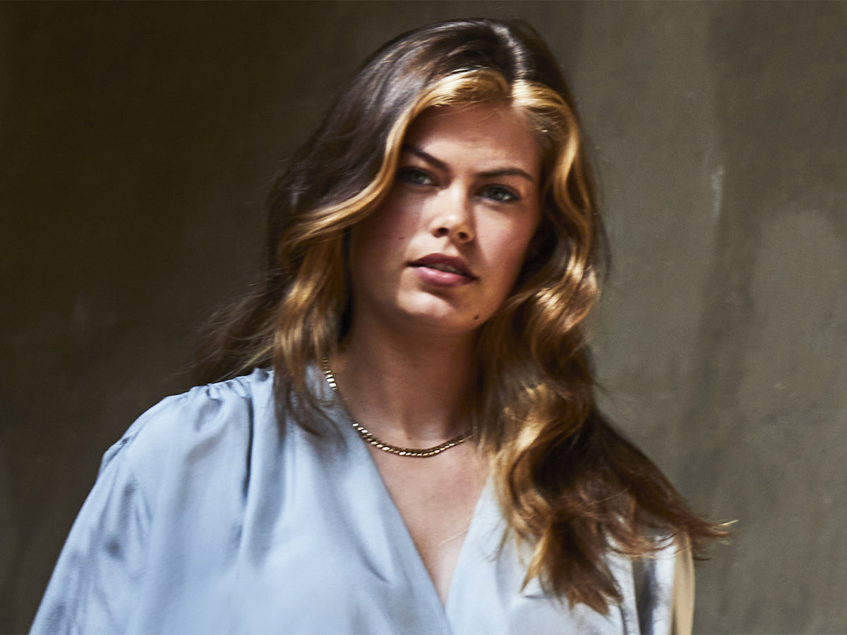 Een vrouw met bruin haar en blonde lokken bij het gezicht voor een groen/bruine achtergrond